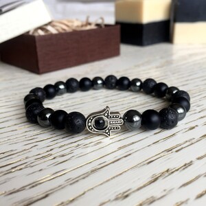 May include: Black beaded bracelet with a silver hamsa hand charm. The beads are made of lava rock and hematite.