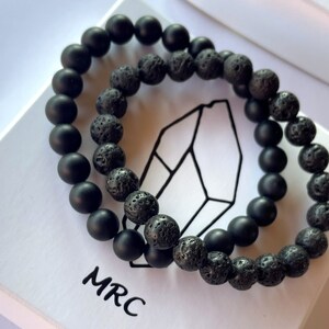 May include: Two black beaded bracelets, one with smooth beads and one with textured beads. The bracelets are on a white surface with the text "MRC" printed on it.