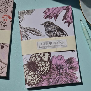 May include: Two small notebooks with floral and bird designs. The notebook on the left has a pink background with brown birds and leaves. The notebook on the right has a white background with a brown butterfly, a pink flower, and a brown bird. Both notebooks have a cream-coloured label with the text "JKEE BOOKS handmade made with love".