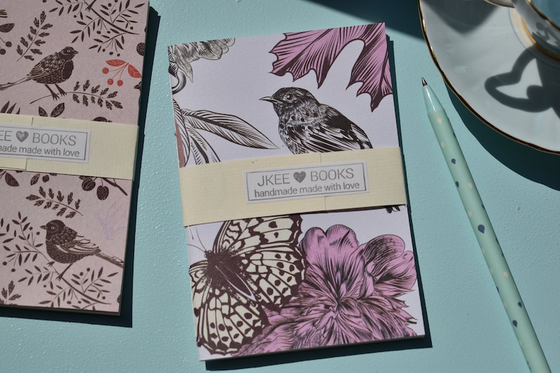 May include: Two small notebooks with floral and bird designs. The notebook on the left has a pink background with brown birds and leaves. The notebook on the right has a white background with a brown butterfly, a pink flower, and a brown bird. Both notebooks have a cream-coloured label with the text "JKEE BOOKS handmade made with love".