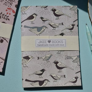 May include: A grey and white notebook with a bird pattern. The notebook has a white label with the text "JKEE BOOKS handmade made with love".
