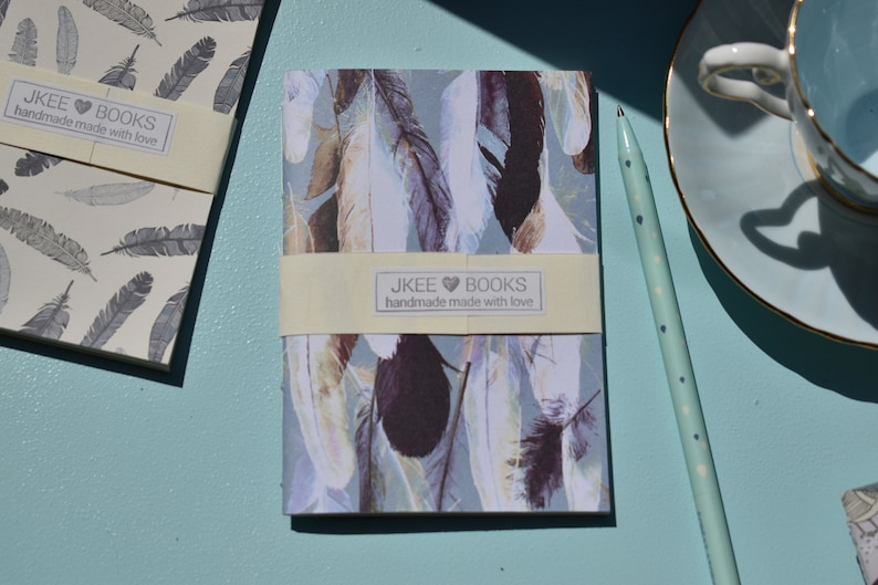 May include: Two notebooks with a feather design. The notebooks are cream and blue with a yellow label that reads "JKEE BOOKS handmade made with love".