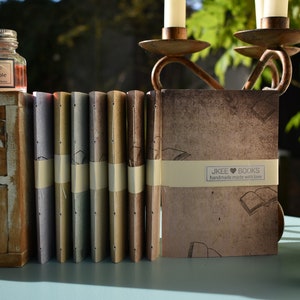 Peut inclure: Une pile de huit petits journaux faits à la main avec des couvertures brunes, beiges et bleues. Les journaux sont empilés sur une table en bois avec une bouteille d'encre et un chandelier en arrière-plan. Les journaux portent une étiquette qui dit "JKEE BOOKS handmade made with love".