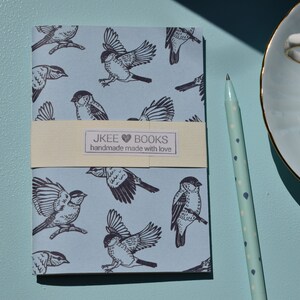 May include: A small blue notebook with a pattern of black birds. The notebook has a cream-coloured label that reads "JKEE &hearts; BOOKS handmade made with love".