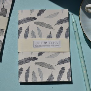 May include: Two small notebooks with a white background and a grey feather pattern. The notebooks have a cream-coloured band with the text "JKEE BOOKS handmade made with love".