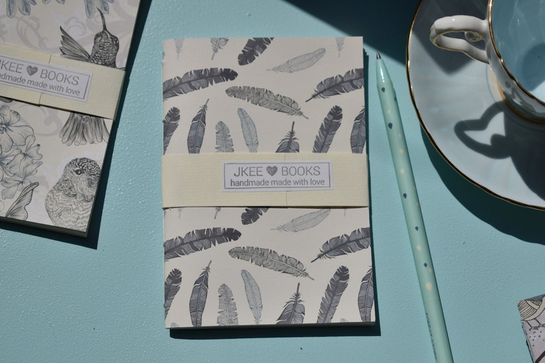 May include: Two small notebooks with a white background and a grey feather pattern. The notebooks have a cream-coloured band with the text "JKEE BOOKS handmade made with love".