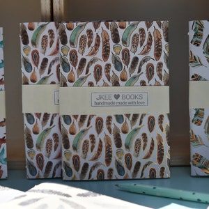 May include: Three small notebooks with different patterned covers. The covers feature a white background with colourful designs of birds and feathers. The notebooks have a label that reads "JKEE BOOKS handmade made with love".