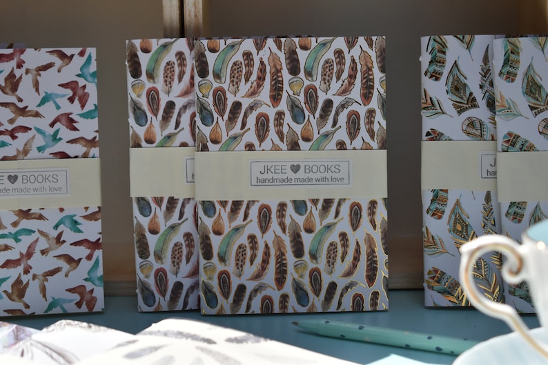 May include: Three small notebooks with different patterned covers. The covers feature a white background with colourful designs of birds and feathers. The notebooks have a label that reads "JKEE BOOKS handmade made with love".