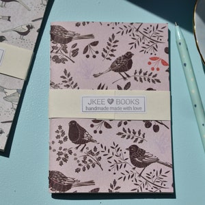 May include: A pink notebook with a floral and bird design. The notebook has a cream-coloured label with the text "JKEE BOOKS handmade made with love".