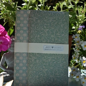Puede incluir: Dos cuadernos con un estampado floral verde claro y crema están envueltos con una banda color crema. La banda presenta el texto "jkee BOOKS handmade made with love". Los cuadernos están rodeados de flores y vegetación.