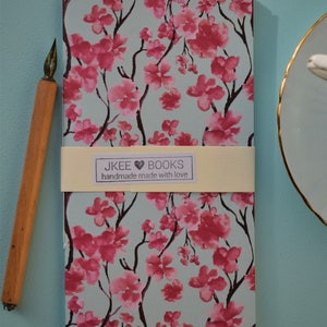 Può includere: Un quaderno blu chiaro con un motivo floreale rosa. Il quaderno ha un'etichetta color crema con il testo "JKEE BOOKS handmade made with love".