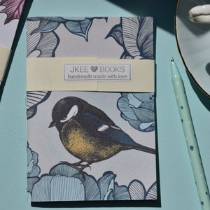 May include: A small, grey notebook with a blue and white floral design and a bird illustration. The notebook has a cream-coloured label that reads "JKEE BOOKS handmade made with love".