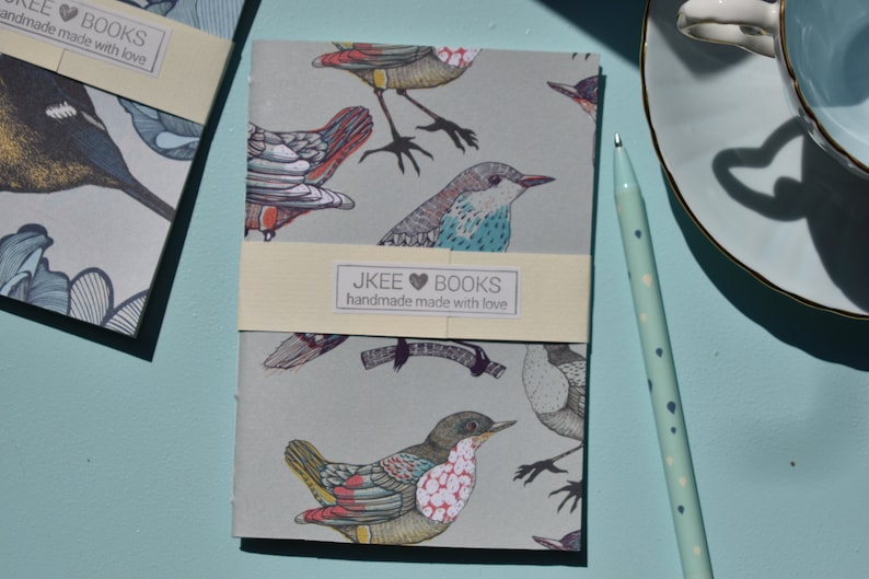 May include: A small, grey notebook with a bird pattern. The cover features colourful birds with intricate details. The notebook has a cream-coloured label that reads "JKEE &hearts; BOOKS handmade with love".