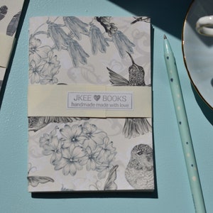 May include: Two small notebooks with floral and feather designs. The notebooks have a cream-coloured cover with a white label that reads "JKEE &hearts; BOOKS handmade made with love".