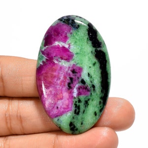 Puede incluir: Piedra preciosa ovalada de color verde y rosa con inclusiones negras. La piedra preciosa tiene una superficie lisa y pulida.