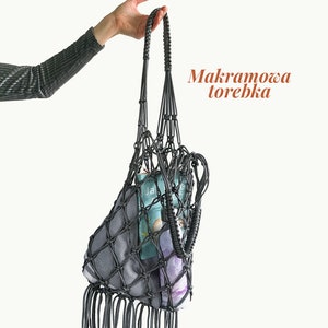Puede incluir: Una bolsa de macramé gris con flecos que cuelgan de la parte inferior. La bolsa está llena de artículos, incluyendo un libro y una botella. El texto "Warsztaty online" y "Makramowa torebka" es visible en la imagen.