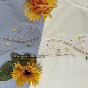 Puede incluir: Dos camisetas de color claro con texto bordado que dice "YOU'VE GOT STARS, THEY'RE IN YOUR EYES" y una imagen estilizada de una luna creciente, estrellas y un cometa. Las camisetas están dispuestas sobre una superficie con flores amarillas.