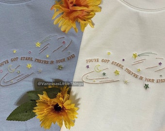 May include: Two light-colored t-shirts with embroidered text that reads "YOU'VE GOT STARS, THEY'RE IN YOUR EYES" and a stylized image of a crescent moon, stars, and a comet. The shirts are laid out on a surface with yellow flowers.