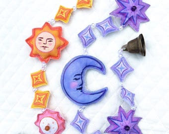 May include: A celestial-themed garland featuring a sun, moon, stars, and bells. The sun is yellow and orange with a smiling face. The moon is blue with a face. Stars are purple. The garland is strung with clear beads.