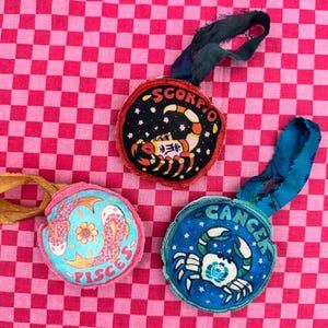 May include: Three fabric ornaments with zodiac signs. A blue ornament with a crab and the word "Cancer", a black ornament with a scorpion and the word "Scorpio", and a blue ornament with two fish and the word "Pisces".