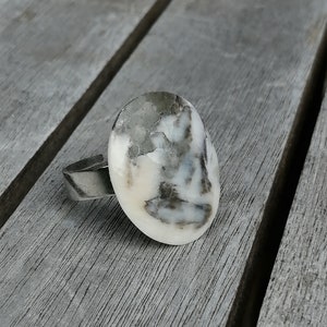 Peut inclure: Une bague en argent avec une pierre ovale blanche et grise sertie au centre. La pierre présente un motif unique de veines grises.