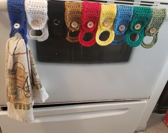 May include: A collection of colorful, crocheted kitchen towel holders in various colors, including blue, white, black, gold, red, yellow, blue, green, and olive green. Each holder has a wooden button closure and is attached to a metal bar on a white oven.