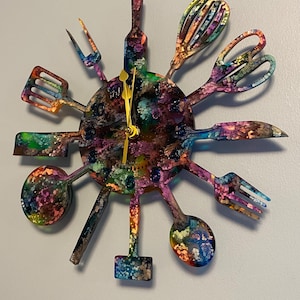 May include: A colorful kitchen-themed wall clock. The clock's face is a circle with various kitchen utensils radiating outwards, including a spatula, whisk, and spoon. The clock hands are gold. The utensils and clock face have a vibrant, abstract paint design.