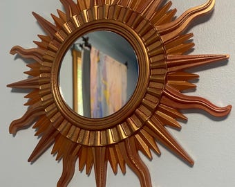 May include: A copper-colored sunburst mirror with a round mirror in the center. The mirror has a decorative design with pointed rays and wavy edges.
