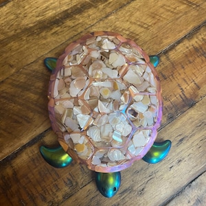 May include: A decorative turtle figurine featuring a shell filled with small, iridescent, light-coloured shell fragments encased in a translucent pink resin frame. The head and four flippers are finished in a metallic, colour-shifting green and blue.