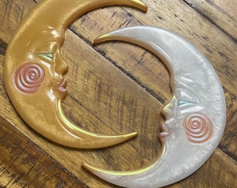 Könnte beinhalten: Zwei halbmondförmige Wanddekorationen mit menschenähnlichen Gesichtern, geschlossenen Augen und spiralförmigen Wangen. Ein Mond hat ein metallisch goldenes Finish, der andere ist perlmuttweiß. Beide Objekte haben glatte, geschwungene Oberflächen.