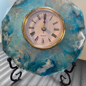 May include: A blue and white resin clock with gold accents and a black metal stand. The clock face has Roman numerals and says "QUARTZ".