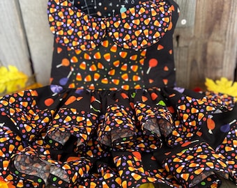 May include: A black Halloween-themed dress with a candy corn pattern. The dress features a ruffled skirt and a Peter Pan collar. The candy corn design includes orange, yellow, and white candy corn pieces, along with purple and orange dots, and red and purple lollipops.