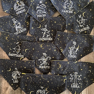 May include: A collection of triangular fabric pet bandanas in black, each featuring a gold celestial pattern of stars and crescent moons. Each bandana displays a unique white line-art zodiac sign and its corresponding name: Aries, Taurus, Gemini, Cancer, Leo, Virgo, Libra, Scorpio, Sagittarius, Capricorn, Aquarius, and Pisces.