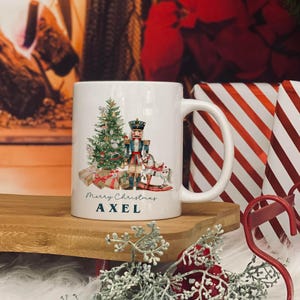 May include: White ceramic mug featuring a Christmas design. The design includes a Christmas tree, a nutcracker, wrapped gifts, and a rocking horse. The text "Merry Christmas AXEL" is printed on the mug.