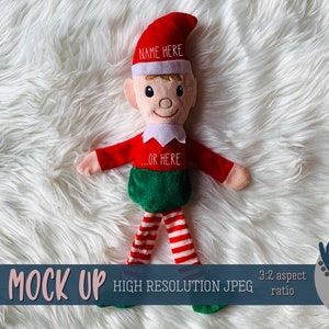May include: A red and green elf doll with a white hat and striped leggings. The elf is lying on a white fluffy surface. The hat has the text "NAME HERE" and the shirt has the text "...OR HERE".