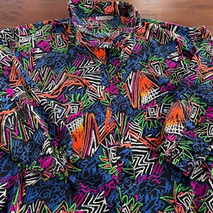 Peut inclure: Une chemise à manches longues noire avec un imprimé géométrique coloré. L'imprimé présente une variété de formes et de lignes dans des tons de rose, orange, vert, bleu et blanc.