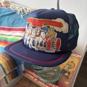 May include: Navy blue trucker hat with a red brim and mesh back. The front panel features an embroidered design of a horse-drawn fire engine with the text "Old Fire Brigade" in red and gold.