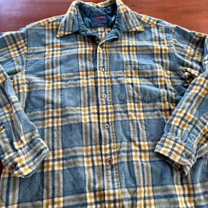 Puede incluir: Una camisa de manga larga con botones y un patrón de cuadros en tonos azul, amarillo y blanco. La camisa tiene un cuello puntiagudo, un bolsillo en el pecho y botones oscuros. Se observa un forro acolchado azul en el interior del cuello.