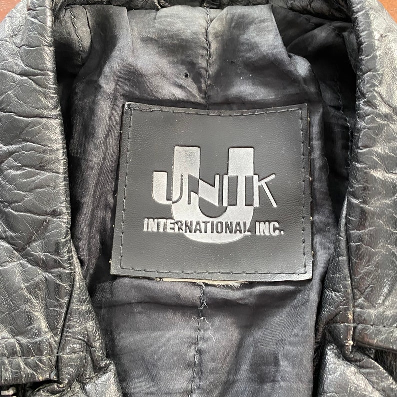 May include: Close-up of a black leather jacket with a black label that reads "UNIK INTERNATIONAL INC."