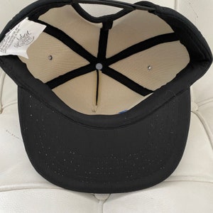 K&ouml;nnte beinhalten: Schwarze Baseballkappe mit wei&szlig;em Netzfutter und schwarzem Riemen. Die Innenseite der Kappe hat ein wei&szlig;es Etikett mit dem Text "Island" in Schwarz.