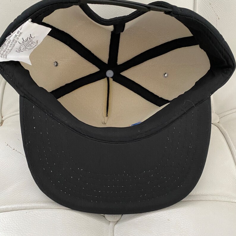 K&ouml;nnte beinhalten: Schwarze Baseballkappe mit wei&szlig;em Netzfutter und schwarzem Riemen. Die Innenseite der Kappe hat ein wei&szlig;es Etikett mit dem Text "Island" in Schwarz.