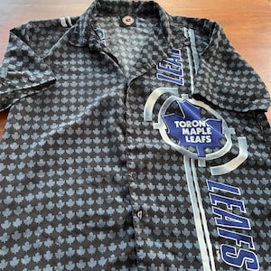Può includere: Camicia nera con bottoni con un motivo di piccole foglie d'acero grigie. La camicia presenta il logo dei Toronto Maple Leafs e la scritta "LEAFS" in bianco e blu. La camicia ha maniche corte.