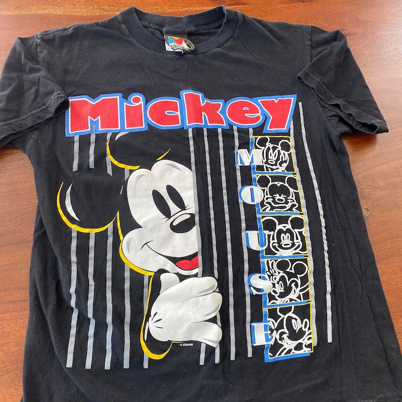 K&ouml;nnte beinhalten: Schwarzes T-Shirt mit einem bunten Motiv von Mickey Mouse, der hinter einer gestreiften Wand hervorlugt. Der Text "Mickey Mouse" ist in roten, blauen und wei&szlig;en Buchstaben gedruckt.