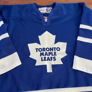 May include: A blue hockey jersey with white stripes on the sleeves and a white Toronto Maple Leafs logo on the front. The logo features a white maple leaf with the words "Toronto Maple Leafs" printed inside.