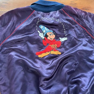May include: Purple satin jacket with a navy blue collar and red trim. Features an embroidered image of Mickey Mouse in a wizard costume, conjuring stars. A vintage-style jacket.