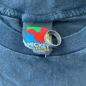 K&ouml;nnte beinhalten: Ein schwarzes T-Shirt mit einem Mickey Mouse Unlimited Multiprint-Etikett. Das Etikett zeigt einen bunten Mickey Mouse-Kopf mit rotem Rand. Das Etikett ist mit einer wei&szlig;en Schnur am Shirt befestigt.