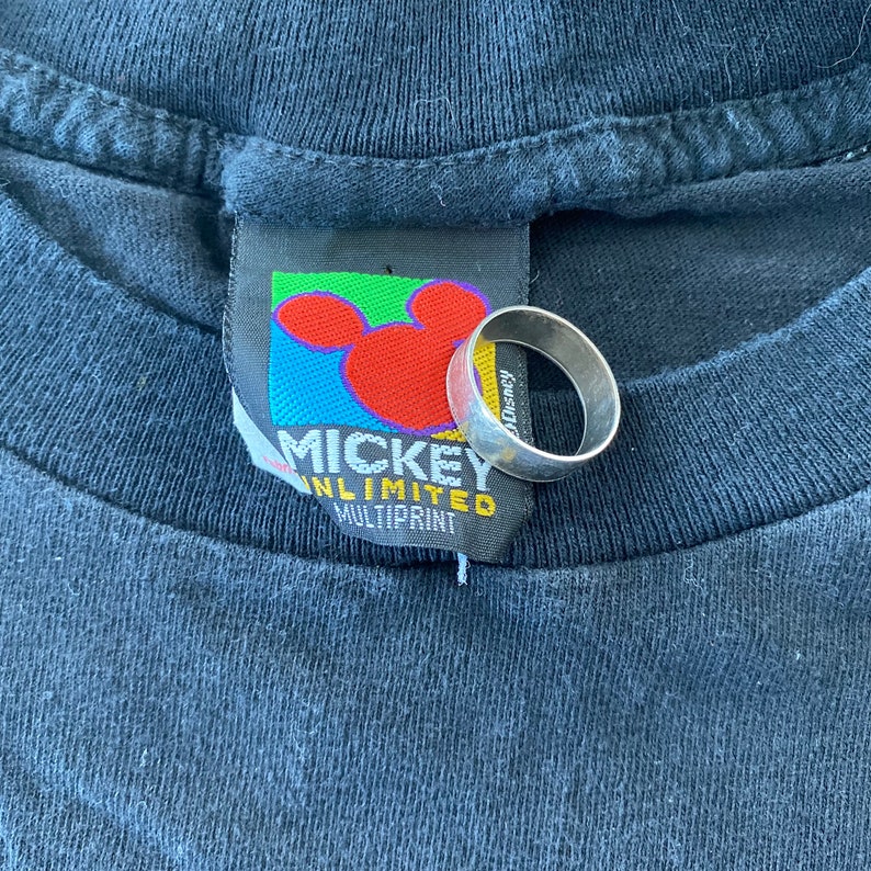 K&ouml;nnte beinhalten: Ein schwarzes T-Shirt mit einem Mickey Mouse Unlimited Multiprint-Etikett. Das Etikett zeigt einen bunten Mickey Mouse-Kopf mit rotem Rand. Das Etikett ist mit einer wei&szlig;en Schnur am Shirt befestigt.