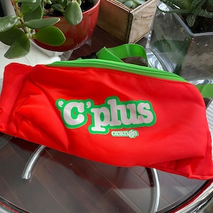 May include: A bright orange fanny pack with a green zipper and strap. The pack has the words "C'plus orange" printed on the front in white and green letters.