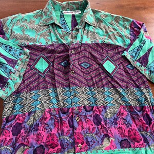 May include: A button-down shirt with a vibrant, patterned design. The shirt features a mix of colors, including teal, purple, pink, and blue, with geometric and floral patterns. The collar and cuffs are teal, and the shirt has a relaxed fit.