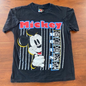 K&ouml;nnte beinhalten: Schwarzes T-Shirt mit einem Motiv von Mickey Mouse, der hinter einer Wand aus vertikalen Streifen hervorlugt. Der Text "Mickey Mouse" ist in roten und blauen Buchstaben &uuml;ber dem Motiv gedruckt.